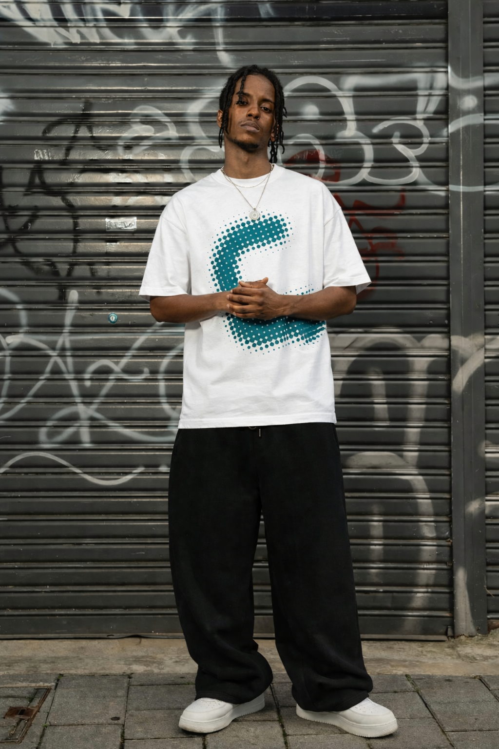 Person wearing a white t-shirt with a blue graphic design, standing in front of a graffitied metal shutter.
